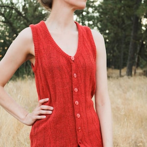 May include: A long red knitted sleeveless vest with a button-up front. The vest is made of a soft, textured fabric and has a relaxed fit.