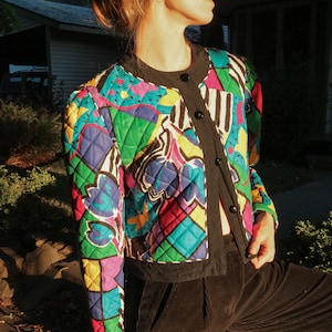 Può includere: Una donna indossa una giacca trapuntata colorata con un motivo geometrico e floreale. La giacca ha un bordo e bottoni neri. La donna indossa pantaloni di velluto a coste neri.