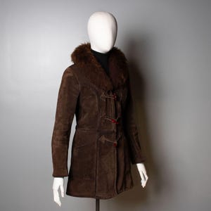 May include: A dark brown suede coat featuring a thick, plush fur collar and a front closure with three toggle buttons. The coat has long sleeves, visible seam detailing along the torso, and a straight hemline, displayed on a white mannequin.