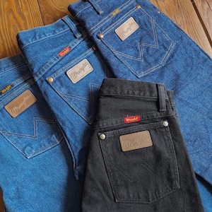 May include: Three pairs of jeans, one black and two blue, with a brown leather Wrangler patch on the back pocket of each.