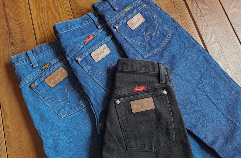 May include: Three pairs of jeans, one black and two blue, with a brown leather Wrangler patch on the back pocket of each.