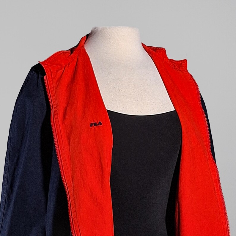 May include: A navy blue and red zip-up jacket with a hood. The jacket has a red interior and a small "FILA" logo on the chest. The jacket is displayed on a mannequin.