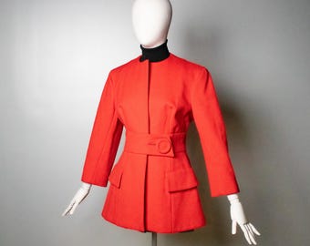 Puede incluir: Una chaqueta de color rojo vibrante con cuello alto, mangas largas y un cinturón estructurado en la cintura con un botón circular grande. La chaqueta incluye dos bolsillos delanteros con solapa y se exhibe en un maniquí con cuello alto negro.