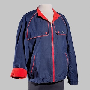 May include: Navy blue jacket with red trim and accents. The jacket features a red collar, cuffs, and zipper. The Fila logo is visible on the right chest. The jacket has two front pockets and a full zipper closure.