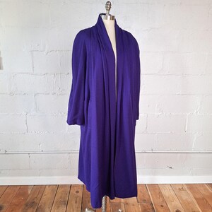 May include: A long, flowing, purple robe with a shawl collar is displayed on a mannequin. The robe has long sleeves and a floor-length hem. The fabric appears to be a soft, draping material.
