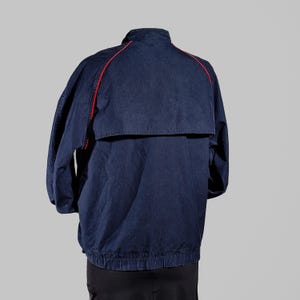 May include: A navy blue jacket with red trim along the shoulders and upper back. The jacket has a high collar and a zippered front. The jacket's design includes a back flap and elastic cuffs and hem. The jacket is made of a durable material.