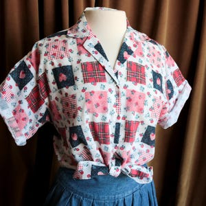 May include: A white short-sleeved button-up shirt with a patchwork design featuring red, pink, and blue squares and hearts. The shirt has a collar and a tie at the waist.
