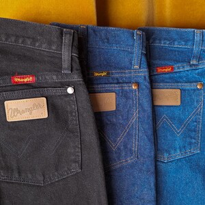 May include: Four pairs of Wrangler jeans in different shades of blue and black. The jeans have a classic five-pocket style with a leather patch on the back pocket that says "Wrangler".