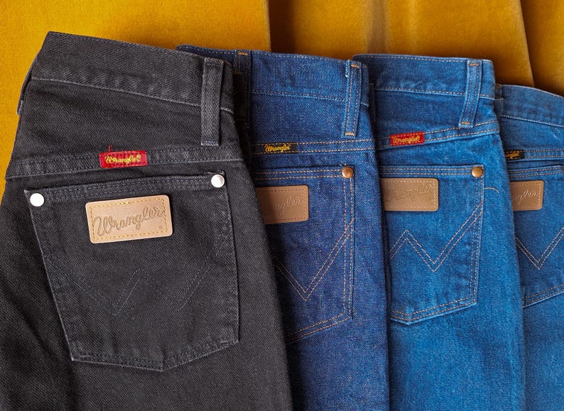 May include: Four pairs of Wrangler jeans in different shades of blue and black. The jeans have a classic five-pocket style with a leather patch on the back pocket that says "Wrangler".