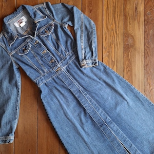 Op de afbeelding: Een lange denim jurk met lange mouwen, een knoopsluiting aan de voorkant en een lange rok. De jurk heeft een klassieke denim look en is een veelzijdig kledingstuk voor elke garderobe.