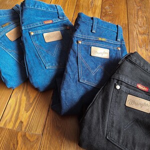 May include: Four pairs of Wrangler jeans in different shades of blue and black. The jeans are all laid out on a wooden surface. The jeans have a classic five-pocket style and a leather patch with the Wrangler logo on the back.