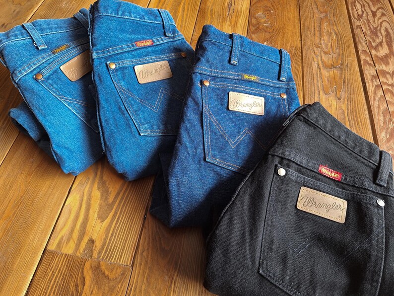 May include: Four pairs of Wrangler jeans in different shades of blue and black. The jeans are all laid out on a wooden surface. The jeans have a classic five-pocket style and a leather patch with the Wrangler logo on the back.