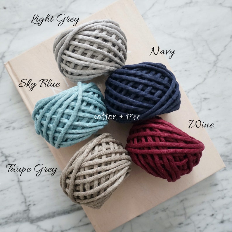 Op de afbeelding: Vijf bollen katoenen touw in verschillende kleuren: lichtgrijs, hemelsblauw, marineblauw, taupegrijs en wijnrood. De bollen zijn in een cirkel op een boek met de tekst "cotton + tree" gerangschikt.