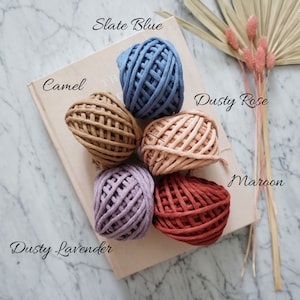Op de afbeelding: Zes bollen katoenen touw in verschillende kleuren, waaronder leisteenblauw, camel, stoffig roze, stoffig lavendel en donkerrood. De bollen zijn in een cirkel op een lichtbruin oppervlak gerangschikt.