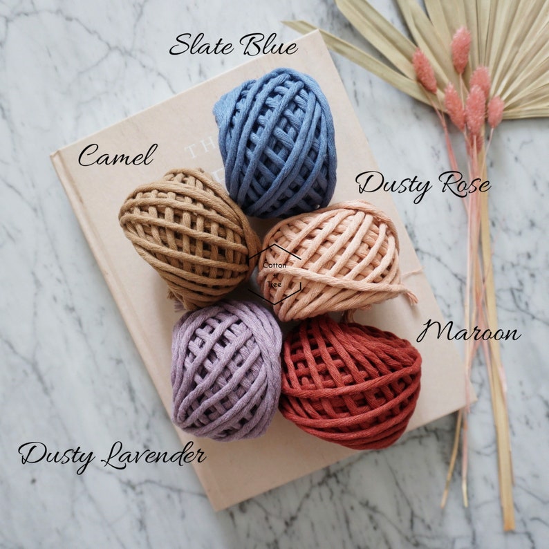 Op de afbeelding: Zes bollen katoenen touw in verschillende kleuren, waaronder leisteenblauw, camel, stoffig roze, stoffig lavendel en donkerrood. De bollen zijn in een cirkel op een lichtbruin oppervlak gerangschikt.