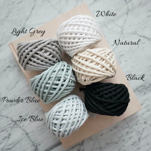 Op de afbeelding: Zes bollen katoenen touw in verschillende kleuren, waaronder wit, lichtgrijs, naturel, poederblauw, ijsblauw en zwart. De bollen zijn in een cirkel op een lichtbruin oppervlak gerangschikt. De tekst "cotton + tree" is zichtbaar in het midden van de afbeelding.