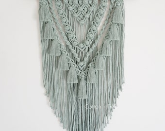 Op de afbeelding: Een stoffig mintgroene macramé wandhanger met een houten lat aan de bovenkant en een franje van kwastjes aan de onderkant. De macramé heeft een geometrisch patroon met een driehoekige vorm. De tekst "Cotton + Tree" is zichtbaar aan de onderkant van de hanger. De tekst "Dusty Mint" is zichtbaar aan de onderkant van de afbeelding.