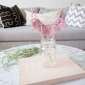 May include: A dusty rose macrame banner is displayed in a clear glass jar with wooden dowels. The banner is placed on a light pink book titled "The Kinfolk Table". The background includes a gray sofa and decorative pillows.