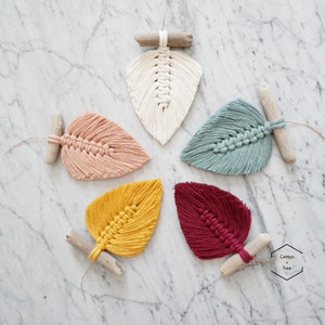 May include: Five macrame leaf decorations in cream, peach, teal, yellow, and burgundy. Each leaf is attached to a piece of driftwood. The leaves are arranged on a marble surface. The Cotton + Tree logo is in the bottom right corner.