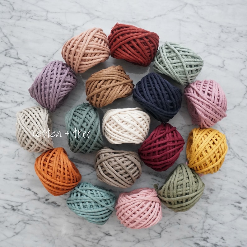 Op de afbeelding: Assortiment van 15 bollen katoenen touw in verschillende kleuren, waaronder rood, blauw, groen, bruin, wit en roze. De bollen zijn in een cirkel op een wit marmeren oppervlak gerangschikt.