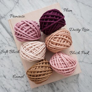 Op de afbeelding: Close-up van acht bollen katoenen touw in verschillende tinten roze, bruin en paars. De bollen zijn gelabeld met de volgende namen: Primrose, Soft Blush, Camel, Blush Pink, Dusty Rose, Plum.