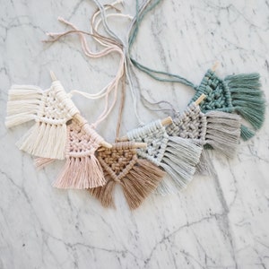 May include: A collection of handmade macrame wall hangings in a variety of pastel colors, including cream, pink, brown, and gray. Each piece features a wooden dowel and a fringed bottom, creating a bohemian aesthetic. The hangings are arranged on a marble surface.