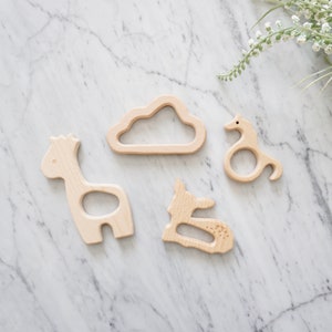 May include: Four wooden baby teethers in the shapes of a giraffe, cloud, seahorse, and deer. The teethers are made of light-colored wood and have smooth edges. The giraffe has a crown, and the deer has dotted markings.
