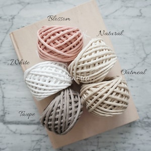 Op de afbeelding: Vijf spoelen katoenen touw in verschillende kleuren: wit, naturel, havermout, taupe en bloesem. De spoelen zijn in een cirkel op een houten oppervlak gerangschikt.