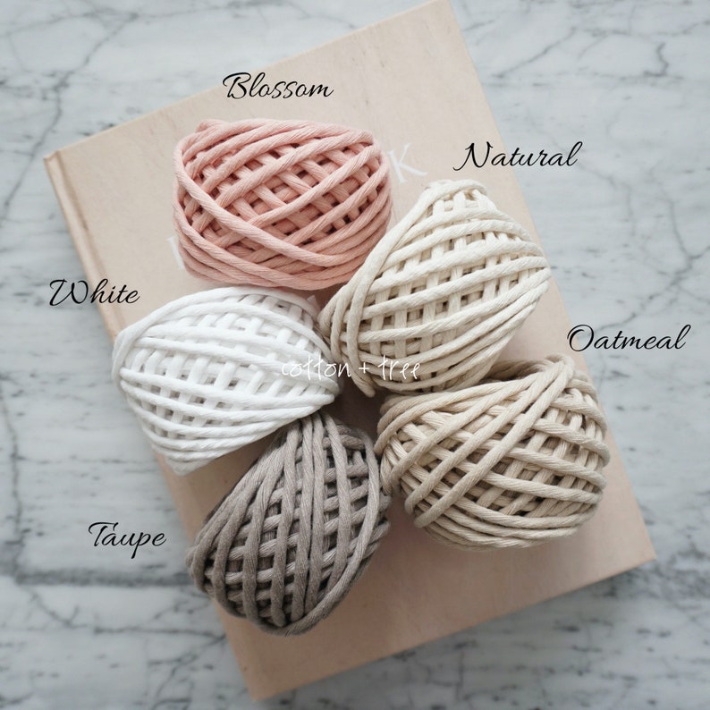 Op de afbeelding: Vijf spoelen katoenen touw in verschillende kleuren: wit, naturel, havermout, taupe en bloesem. De spoelen zijn in een cirkel op een houten oppervlak gerangschikt.