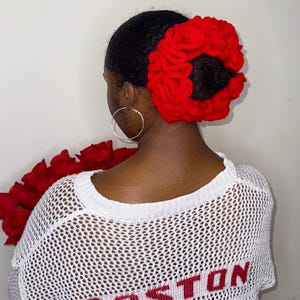 May include: A woman's hair is styled with a large, vibrant red crocheted hair accessory. She wears a white, open-knit sweater with the word "BOSTON" in red letters across the back. Silver hoop earrings complete the look.