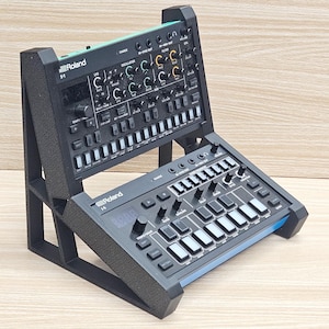 May include: Black Roland S-1 and H-1 synthesizers on a black stand. The S-1 is angled upward, displaying various knobs and buttons. The H-1 is positioned below, featuring a keyboard and a digital display. The stand provides support and an elevated view.