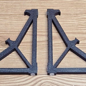 May include: Two dark gray metal speaker stands. Each stand has a triangular design with a top support and a base with small rubber feet. The stands are designed to elevate speakers.