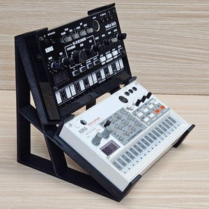 May include: A black synthesizer stand holding two Korg Volca synthesizers. The top synthesizer is black with white text and knobs, labeled "MS-20 Resonator" and "volca kick". The bottom synthesizer is white with gray knobs and buttons, labeled "volca sample".