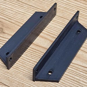 May include: Two black metal shelf brackets with a right-angle design. Each bracket has a flat, rectangular base with pre-drilled holes for mounting. The brackets are made of a dark, matte material and are positioned on a light wood surface.