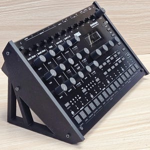 May include: A black Korg drum machine, angled on a black stand. The machine has numerous knobs, buttons, and a small display screen. The text "drumlogue" is visible on the top right. The stand is a simple, geometric design.