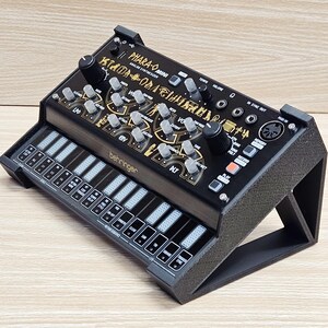 May include: Black analog synthesizer with a mini keyboard. The synthesizer has knobs, buttons, and the text "PHARA-O mini" and "behringer" in gold lettering. The keyboard has black and white keys.