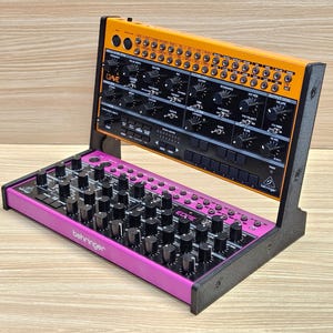 May include: A synthesizer with an orange top panel and a pink bottom panel. The synthesizer has numerous knobs and buttons. The word "DAVE" is visible on the top panel, and "EDGE" is on the bottom panel. The brand name "behringer" is also visible.