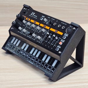 May include: A black Behringer JT Mini analog synthesizer with a built-in keyboard. The synthesizer has various knobs, sliders, and buttons labeled LFO, VCO, VCF, and ENV. The keyboard has white and black keys, and the device is mounted on a textured black stand.
