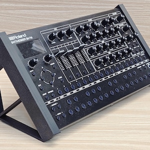 May include: Black synthesizer with numerous knobs, sliders, and buttons. The text "Roland Synthesizer SH-01a" is visible on the top left. The device is angled on a black stand, showcasing its controls and design.