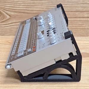 May include: A silver and gray TR-06 Drum machine, angled on a black stand. The drum machine has numerous knobs and buttons. The stand is made of black plastic and has an arched design.