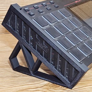 May include: A black Akai MPC Live music production center resting on a black 3D-printed stand. The MPC Live features numerous gray pads, buttons, and a screen. The stand is angled, providing an ergonomic setup for music production.