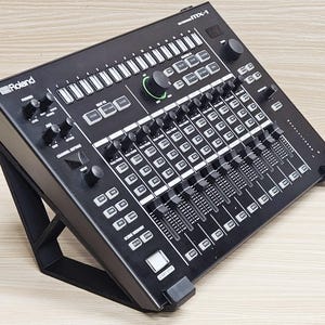 May include: Black Roland MX-1 mixer on a black stand. The mixer features numerous knobs, faders, and buttons, with white labels and a green control knob. The Roland logo is visible on the top left corner. The stand is angled, providing an elevated view of the mixer.