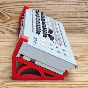 May include: A gray TR-09 drum machine with black knobs and buttons, resting on a red stand. The stand is angled, providing support for the electronic device. The Roland logo is visible on the device.