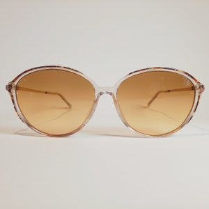 Puede incluir: Gafas de sol de gran tamaño con montura transparente y lentes degradadas marrones. La parte superior de la montura presenta un patrón decorativo. Las gafas tienen finas varillas doradas y un diseño retro.