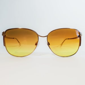 Puede incluir: Gafas de sol doradas con lentes degradadas, que van del amarillo en la parte inferior al ámbar en la parte superior. La montura tiene un diseño delicado, similar a un alambre, con un sutil acento rectangular en las patillas. Las lentes son ovaladas.