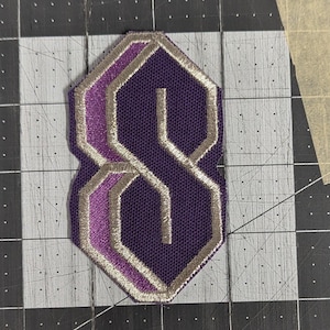 May include: Purple and silver embroidered patch in the shape of the letter "S".