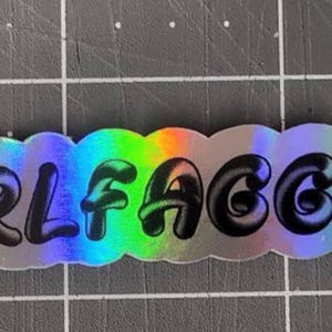 May include: A silver sticker with a rainbow iridescent background. The sticker has the text "GIRL FAGGOT" in black, bubble-like letters.