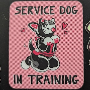 Puede incluir: Un parche rosa y blanco para perros de servicio en entrenamiento con un perro de dibujos animados en blanco y negro que lleva un chaleco rojo y un hueso en la boca. El texto "SERVICE DOG IN TRAINING" está impreso en el parche.