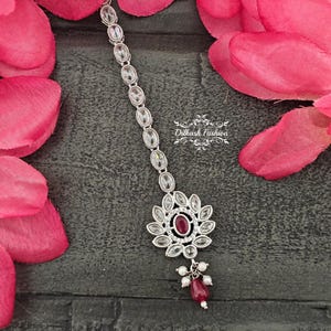 May include: Silver-toned headpiece with a central flower-shaped design featuring a ruby-colored stone. The headpiece has a series of oval-shaped clear stones and a dangling cluster of pearls and a ruby-colored bead. The background includes pink flower petals.