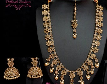 May include: A gold necklace set with a long chain, a matching tikka, and earrings. The necklace features a design of small, round, clear stones set in a floral pattern. The set is accented with small, white pearls.